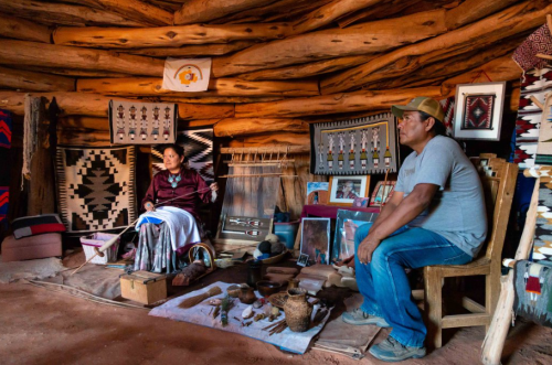 Two individuals sit inside a traditional dwelling, surrounded by handmade crafts and woven textiles.