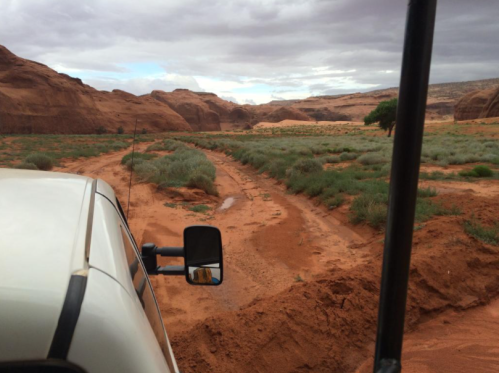 A view from a vehicle showing a sandy path through a desert landscape with sparse vegetation and rocky formations.