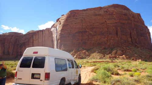 A white van parked near a towering red rock formation under a blue sky with scattered clouds.
