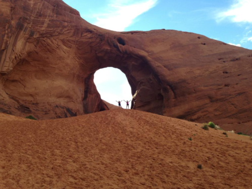 Silhouetted figures stand in a large rock arch against a blue sky, surrounded by sandy terrain.