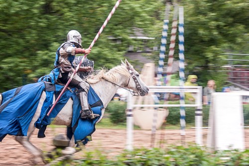 A knight in armor rides a white horse, holding a lance, during a jousting event in a lush outdoor setting.
