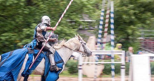 A knight in armor rides a white horse, wielding a lance, during a jousting event in a lush outdoor setting.