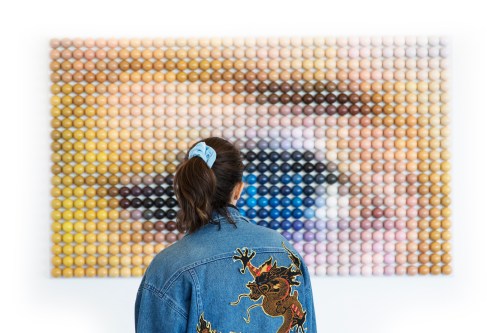 A person in a denim jacket stands in front of a colorful eye artwork made of circular patterns.