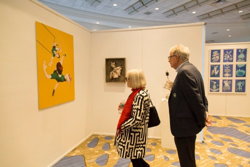 Two visitors observe artwork in a gallery, one wearing a patterned coat and the other holding a drink.