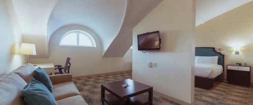 Cozy hotel room with a sloped ceiling, featuring a sofa, desk, TV, and a bed with soft lighting.