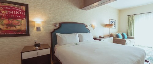 A cozy hotel room featuring a queen bed, bedside tables, and a seating area with natural light from a window.