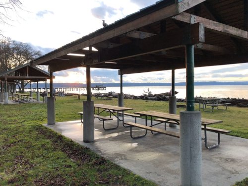 A picnic area with tables under a shelter, overlooking a calm waterway and a distant pier at sunset.