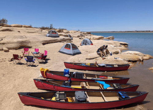 Canoes on a sandy shore with tents and camping chairs nearby, set against a calm lake and clear blue sky.