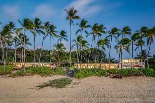 A beach scene at dusk with palm trees, sandy shore, and a building in the background surrounded by lush greenery.