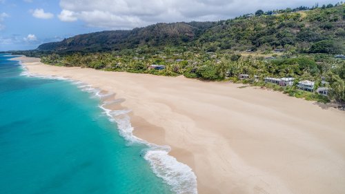 Aerial view of a serene beach with turquoise water, soft sand, and lush green hills in the background.