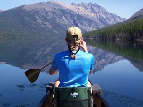 A person in a blue shirt paddles a canoe on a calm lake, surrounded by mountains and trees under a clear sky.
