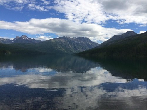 A serene lake reflects mountains and a cloudy sky, creating a tranquil natural landscape.