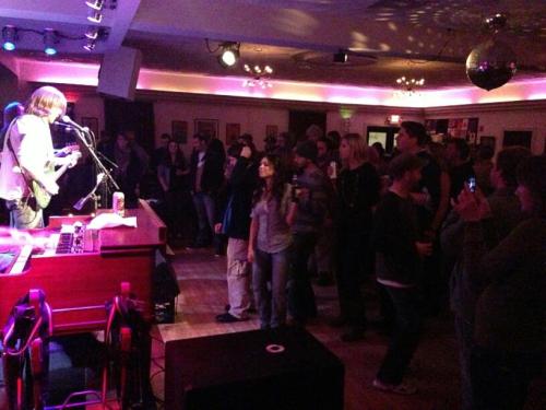 A lively crowd enjoys a live music performance in a dimly lit venue with colorful lights and a disco ball.