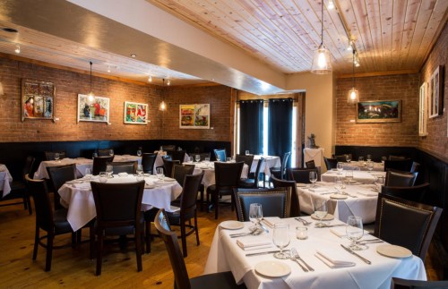 Cozy restaurant interior with wooden ceiling, brick walls, and neatly set tables with white tablecloths.