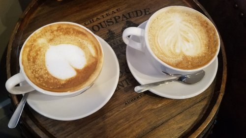 Two cups of coffee with latte art, resting on a wooden surface. One cup has a heart design, the other a swirl.