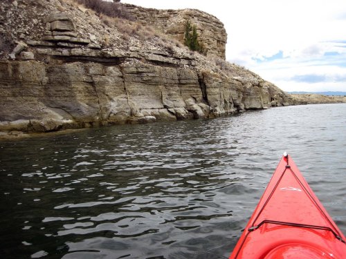 A red kayak approaches rocky cliffs along a calm waterway under a cloudy sky.