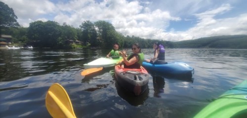 Three people kayaking on a calm lake surrounded by trees under a partly cloudy sky.
