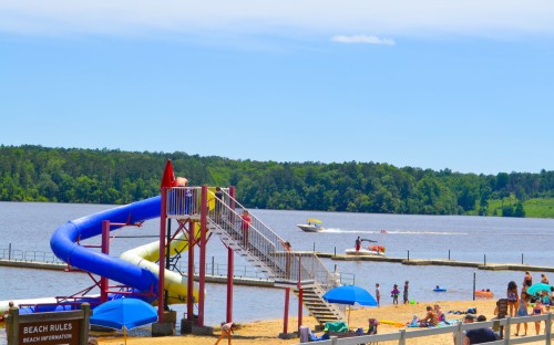 A colorful water slide at a beach with people sunbathing, swimming, and a boat on the lake under a clear blue sky.
