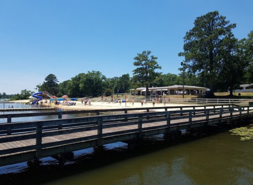 A sunny beach scene with people enjoying the water, a playground, and a wooden pier in the foreground.