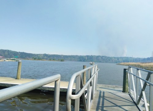 A view of a calm lake from a wooden dock, with distant hills and smoke rising in the background.