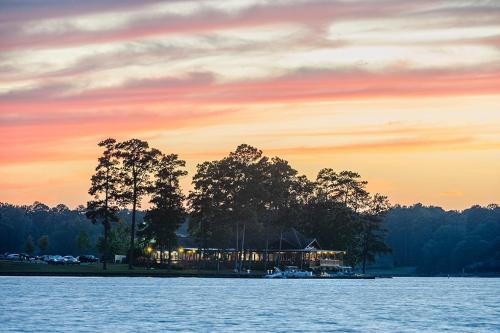 A serene lakeside view at sunset, featuring trees and a building illuminated by warm lights against a colorful sky.