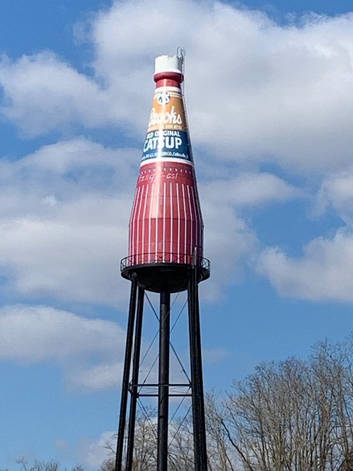 A large water tower shaped like a ketchup bottle, painted red and white, against a blue sky with clouds.
