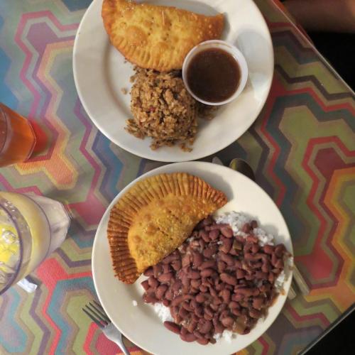 Two plates of food featuring rice, beans, empanadas, and a dipping sauce, set on a colorful patterned table.