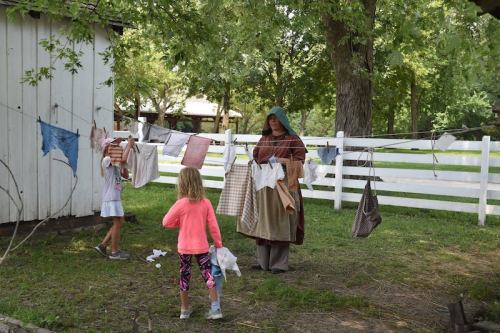 A woman hangs laundry on a line while a child observes in a grassy area with trees and a white fence.