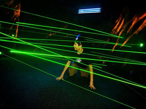 A person crouches on the ground, navigating through bright green laser beams in a dark environment.