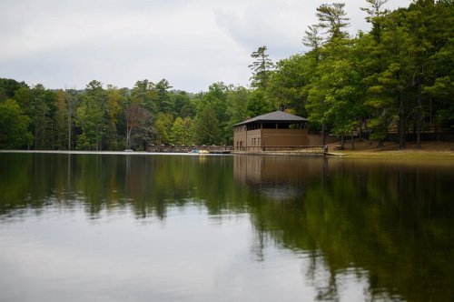 A serene lake scene with a wooden building and lush green trees reflected in the calm water under a cloudy sky.