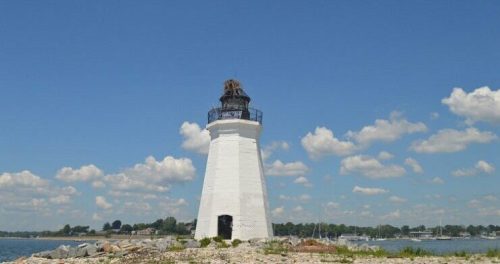 A white lighthouse stands on a rocky shore under a blue sky with fluffy clouds, overlooking a calm body of water.