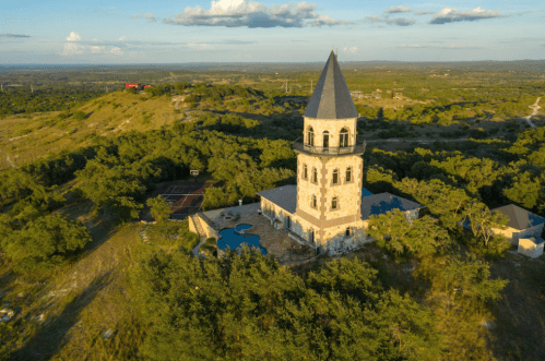 Aerial view of a tall stone tower surrounded by greenery and a pool, set against a hilly landscape under a blue sky.