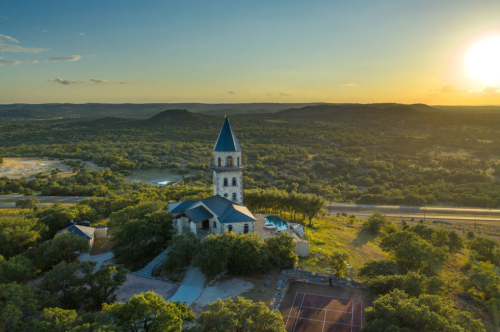 Aerial view of a stone church with a tall spire, surrounded by greenery and hills at sunset.