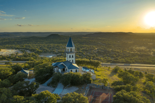 Aerial view of a stone church with a tall spire, surrounded by greenery and hills at sunset.