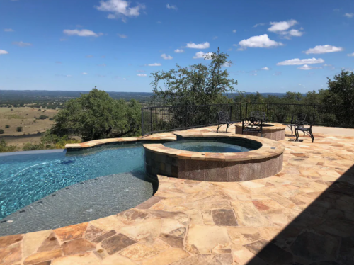 A scenic pool area with a hot tub, surrounded by stone, overlooking a vast landscape under a blue sky.