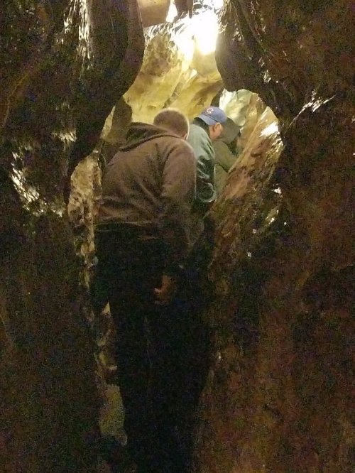 Two people navigating a narrow, rocky cave passage illuminated by overhead lights.