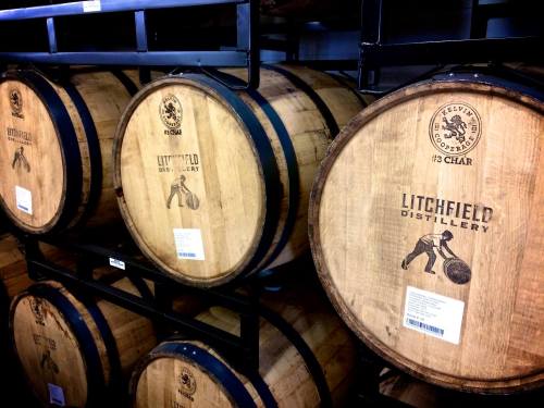Wooden barrels stacked on shelves, labeled with "Litchfield Distillery" and a logo featuring a person rolling a barrel.