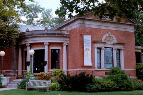 Historic brick building with a rounded entrance, surrounded by greenery and a bench in front.