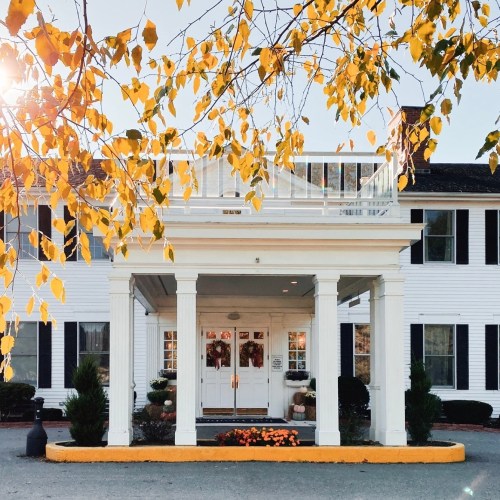 A white building with a porch, surrounded by autumn leaves and flowers, under a bright sun.