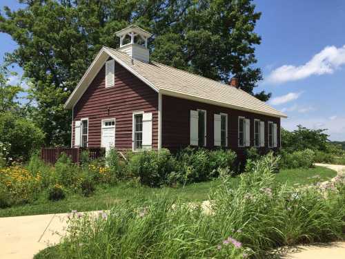 A red wooden schoolhouse with white trim, surrounded by greenery and flowers, under a blue sky with scattered clouds.