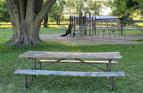 A weathered picnic table in a grassy area near a playground with climbing structures and trees in the background.