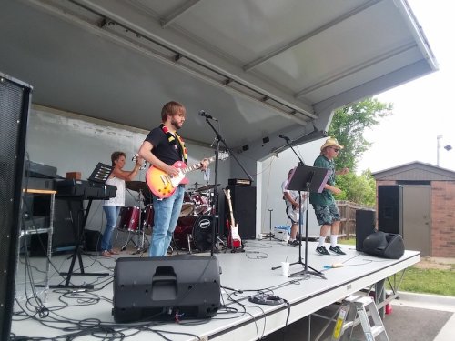 A band performs on stage, featuring a guitarist, drummer, and vocalist, with instruments and equipment visible.