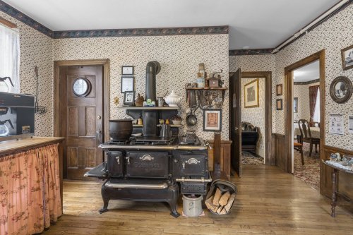 A vintage kitchen featuring a black stove, wooden cabinets, floral wallpaper, and a doorway leading to another room.