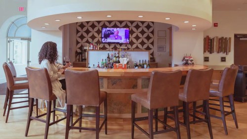 A woman sits at a bar, sipping wine, with a TV screen and various bottles in the background.