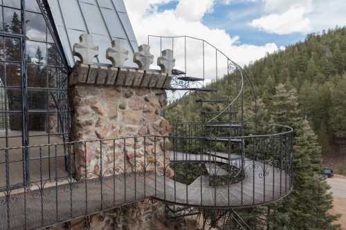 A spiral staircase leads to a balcony on a stone and glass building, surrounded by trees and a cloudy sky.