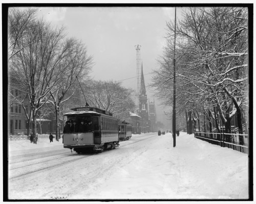 A snowy street scene featuring a tram, trees covered in snow, and a church steeple in the background.