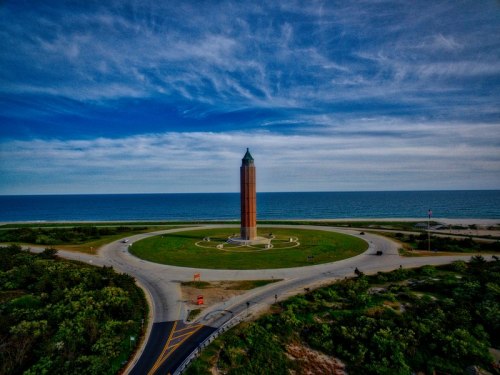 A tall, cylindrical tower stands in a circular park by the ocean, surrounded by greenery and a winding road.