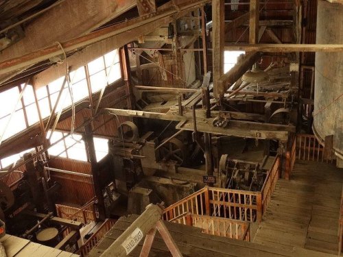 Interior view of an old industrial facility with machinery, wooden beams, and large windows. Dusty and rustic atmosphere.