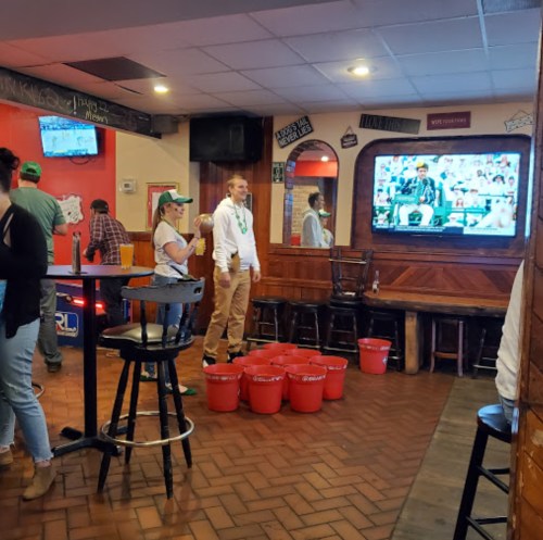 A lively bar scene with people playing beer pong and watching a game on TV, decorated for a festive atmosphere.