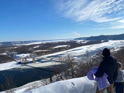 A person and child overlook a snowy landscape with a river and distant hills under a clear blue sky.